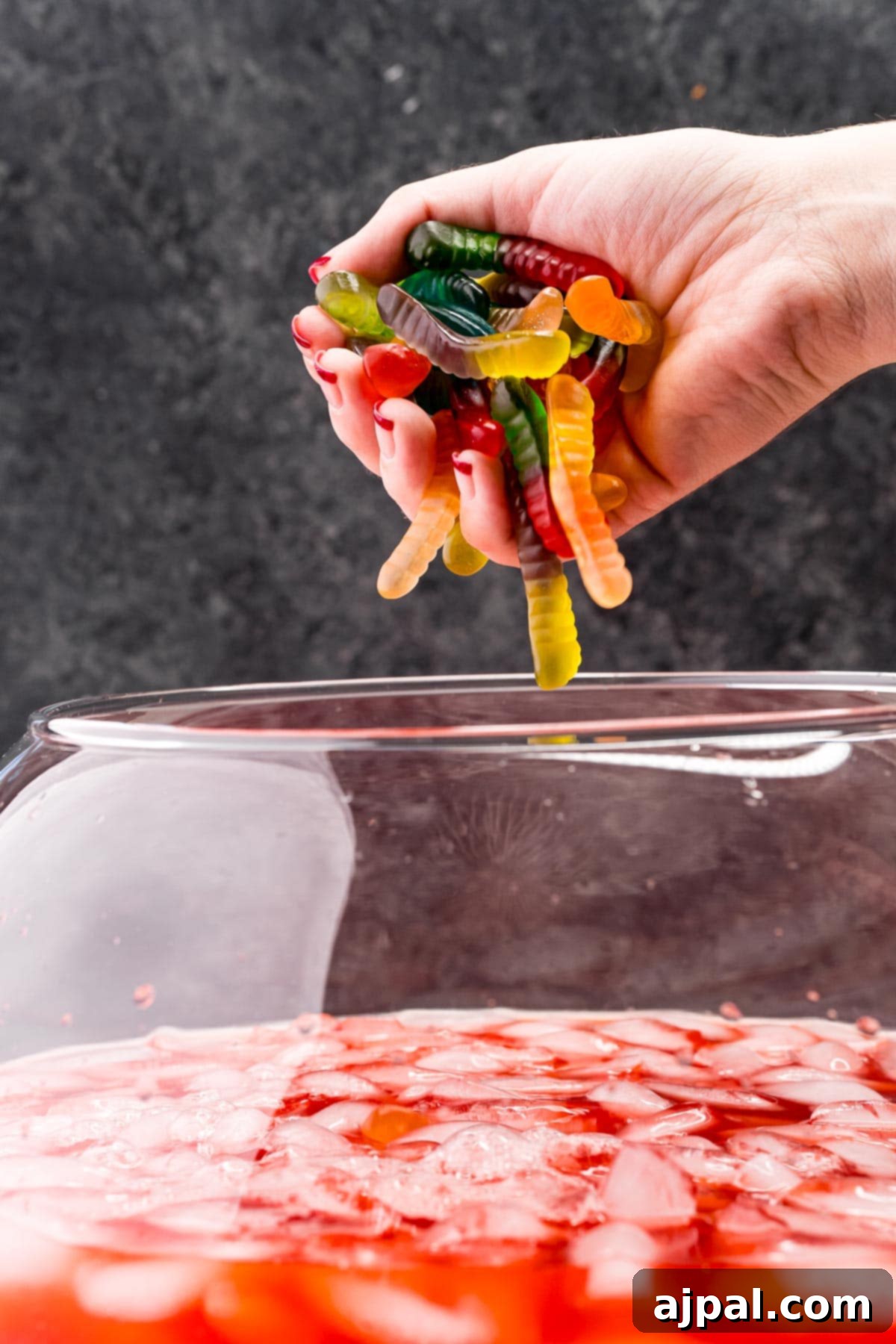 Gummy worms are being dropped into the vibrant red punch, ready to add a creepy-crawly touch.