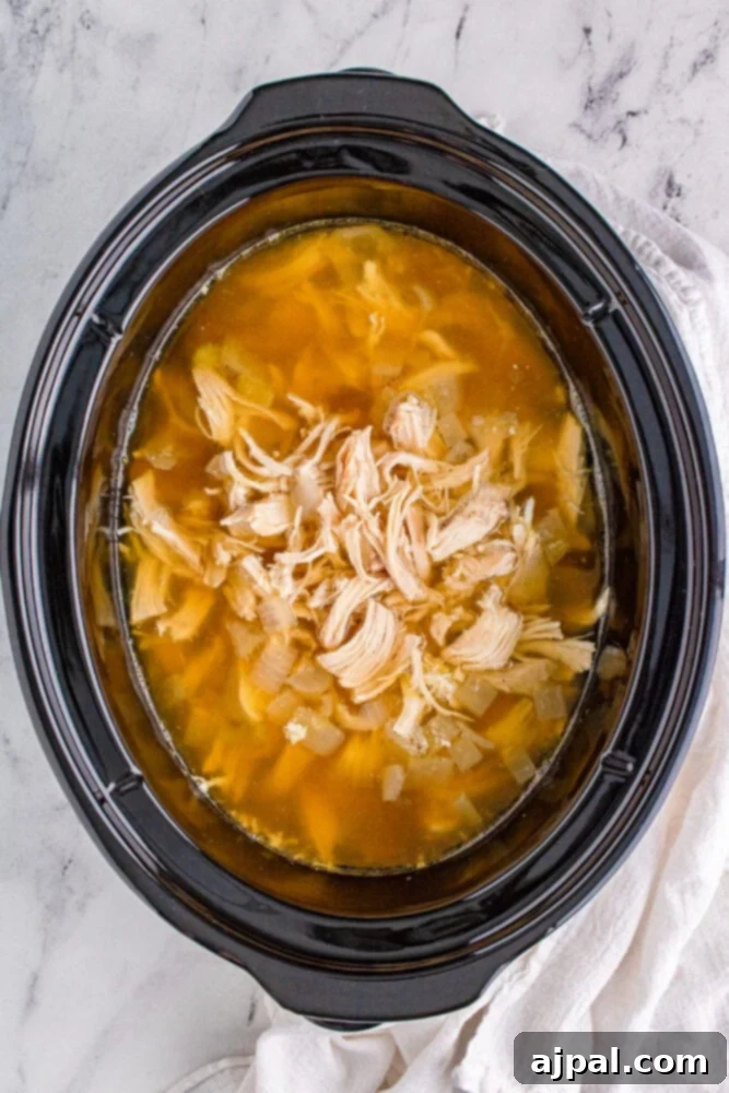 Cooked chicken shredded into pieces and submerged in a flavorful broth with vegetables in a slow cooker.