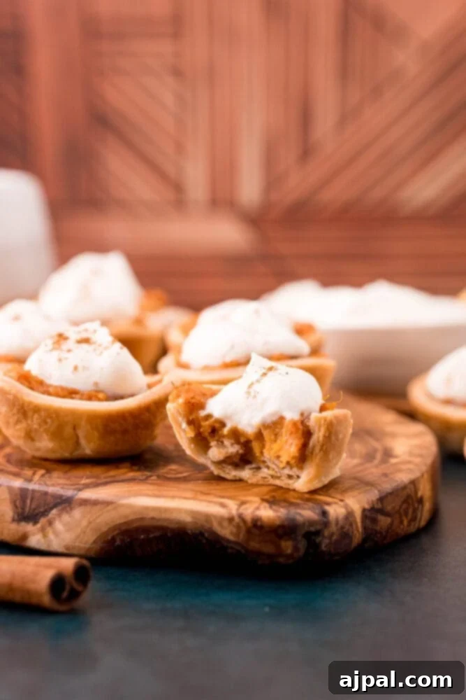 Three mini sweet potato pies, adorned with fluffy whipped cream, sit on a wooden cutting board with a small spoon, showcasing a delightful and easy dessert.