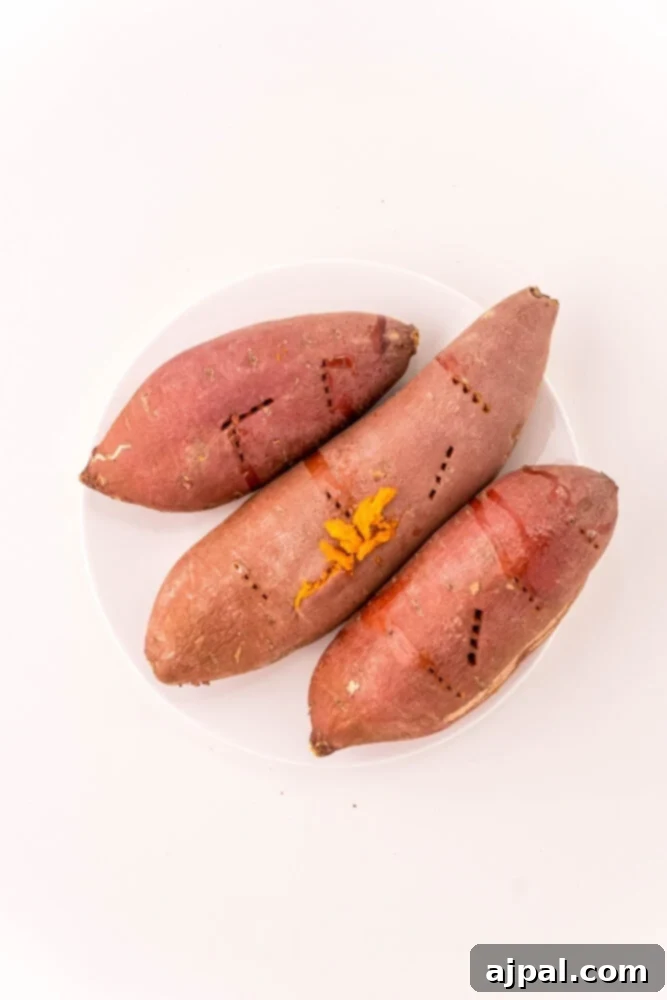 Three perfectly roasted sweet potatoes with golden-brown skins, resting on a rustic wooden board, ready for mashing.