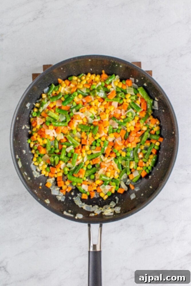 Frozen mixed vegetables added to a pan with sautéed onions, cooking on the stovetop.