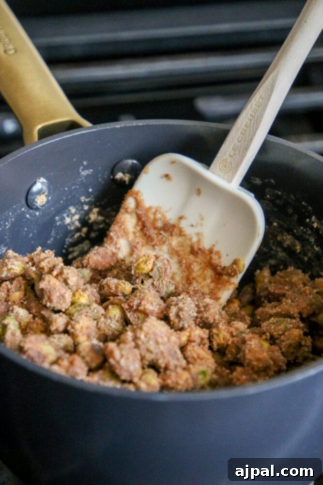 A spatula gently stirring pistachios into a glossy, amber-colored candy mixture in a saucepan, ensuring an even coating.
