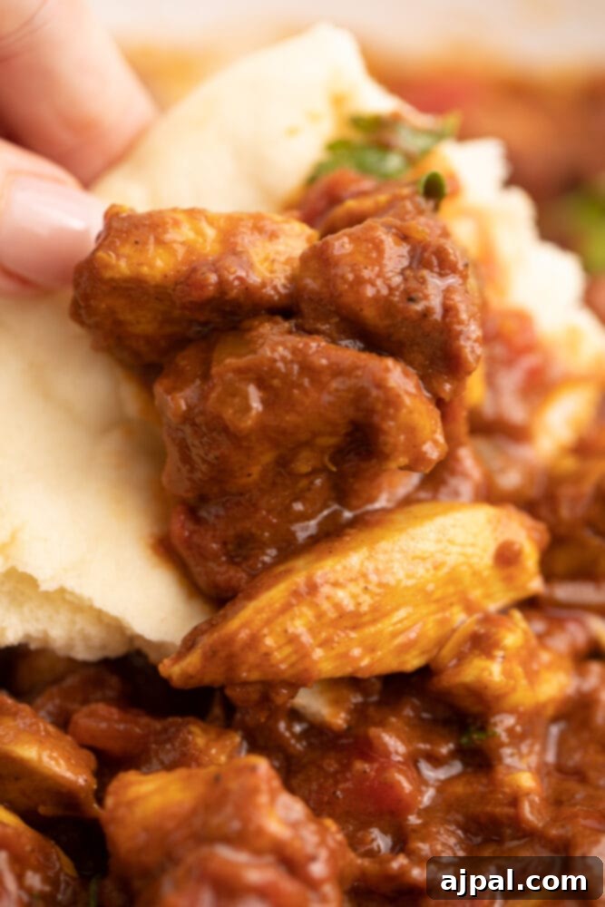 A hand expertly scoops a portion of hot Chicken Madras Curry with fluffy naan bread, showcasing the meal's enticing texture and richness.