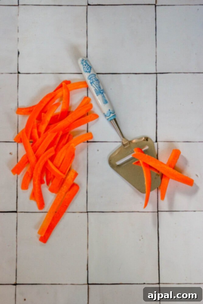 Thinly sliced carrots on a cutting board next to a vegetable peeler, illustrating the preparation step for carrot bacon.