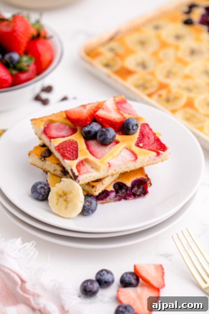 A beautifully stacked plate of golden sheet pan pancakes adorned with fresh strawberries, blueberries, and sliced bananas, ready for a delicious breakfast.