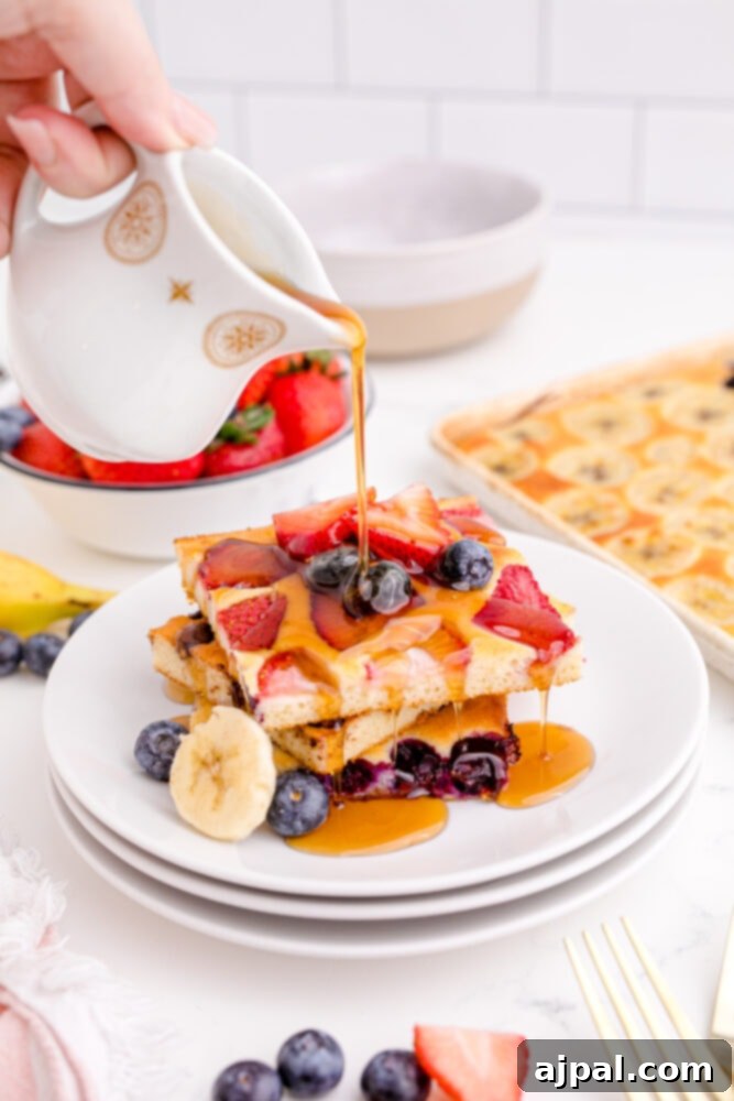 A hand gently pouring a stream of rich maple syrup over a generous stack of sheet pan pancakes, artfully topped with an assortment of fresh berries and sliced fruit, promising a sweet and satisfying breakfast.