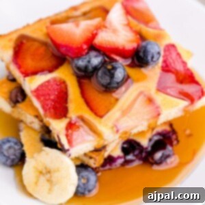A top-down view of a stack of golden sheet pan pancakes on a white plate, artfully garnished with a mix of fresh blueberries and raspberries, and drizzled with syrup, ready to be enjoyed for breakfast.