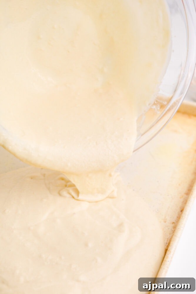 Pancake batter being carefully poured from a bowl into a large baking sheet, illustrating the first step in making sheet pan pancakes.