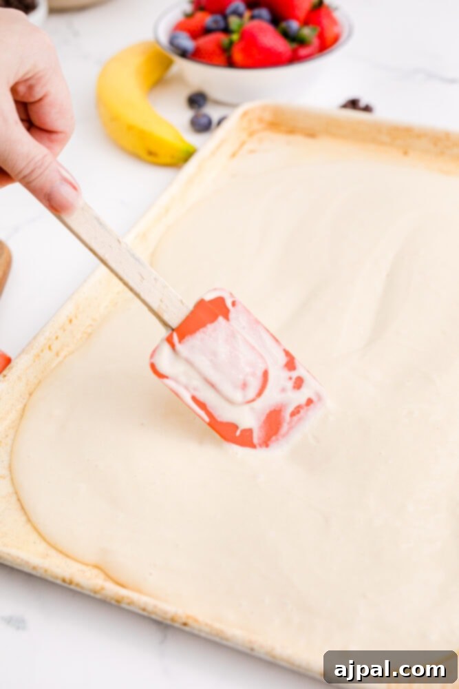 A spatula smoothly spreading a layer of pancake batter evenly across a sheet pan, ensuring a uniform thickness for baking.