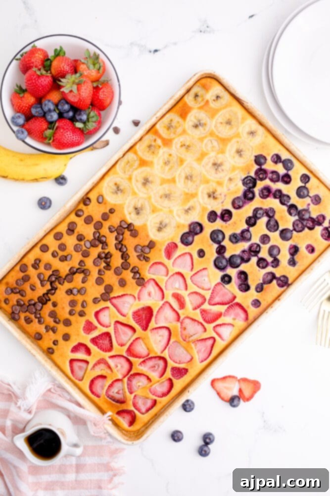 A sheet pan filled with perfectly baked pancakes, divided into sections and topped with a colorful array of fresh fruit and chocolate chips, accompanied by a bowl of assorted berries.