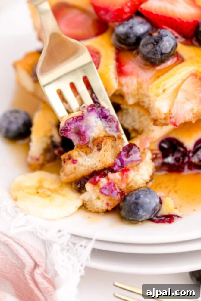 A fork lifts a fluffy piece of pancake from a stack, which is garnished with blueberries, bananas, and a generous drizzle of syrup, highlighting the delicious texture and presentation.