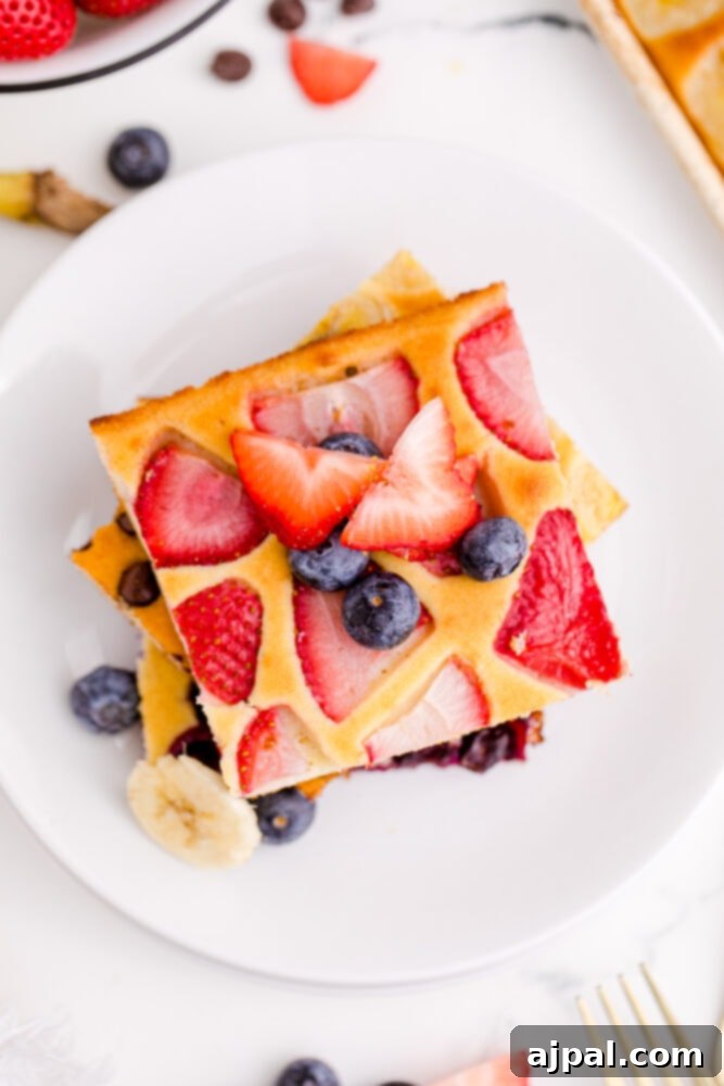 An overhead shot showcasing a beautifully arranged stack of fluffy sheet pan pancakes on a white plate, garnished with an abundance of fresh strawberries and blueberries.
