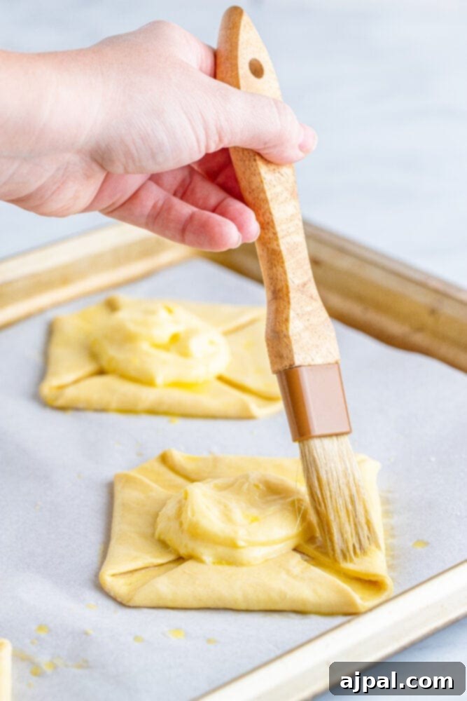 A hand brushing egg wash onto the edges of a puff pastry square on a baking sheet.