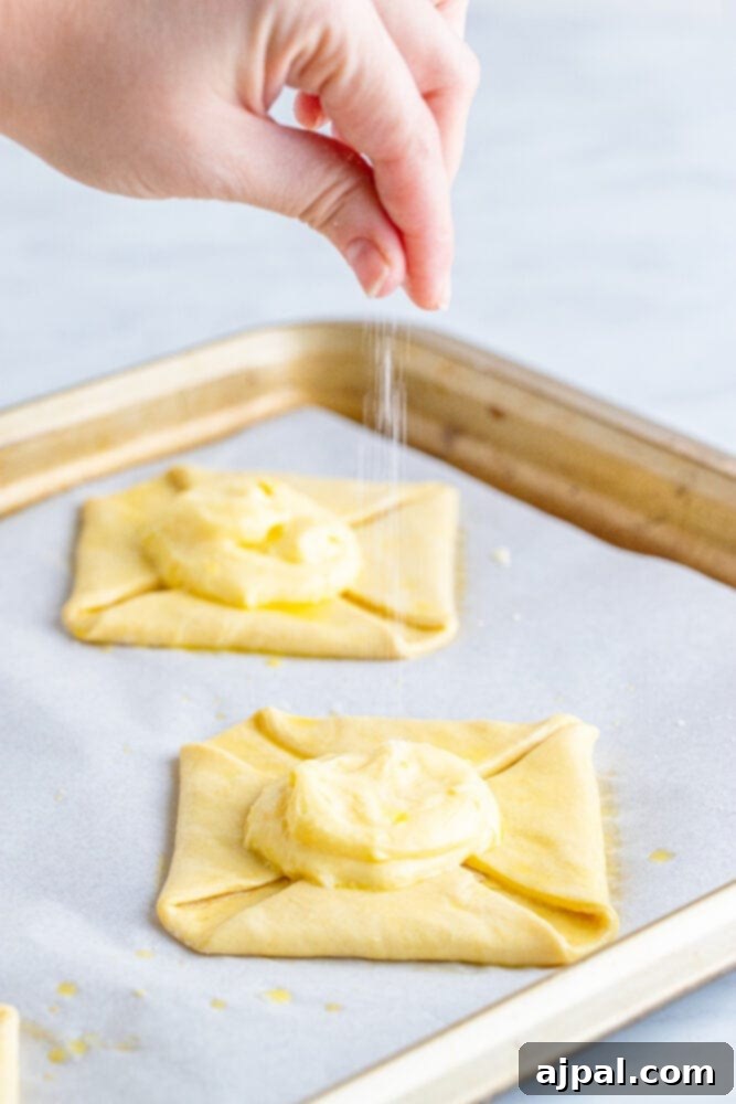 A hand sprinkling sugar over a cheese danish on a baking sheet, adding a sweet crunch.