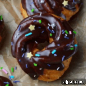 A close-up of a cruller donut covered in rich chocolate glaze and decorated with star-shaped, multicolored sprinkles. The cruller donut rests on brown parchment paper, highlighting its delicious detail.