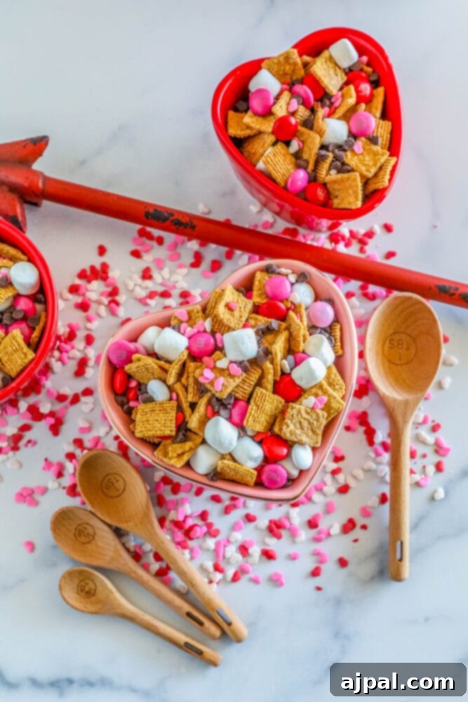 Valentine Smores Snack Mix 3 Heart-shaped bowl filled with Valentine's S'mores Snack Mix, surrounded by wooden spoons, highlighting the delightful combination of marshmallows, pink M&M's, sprinkles, and chocolate chips.