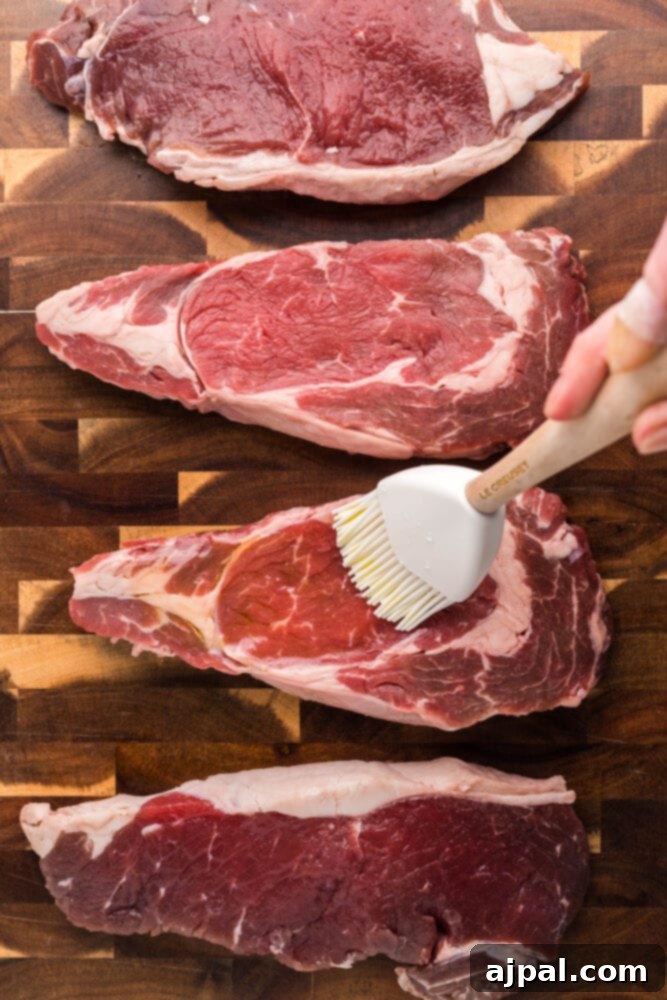 A basting brush applying olive oil to a raw New York Strip steak on a cutting board.