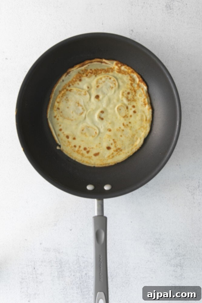 A thin crepe cooking in a frying pan on a white surface.