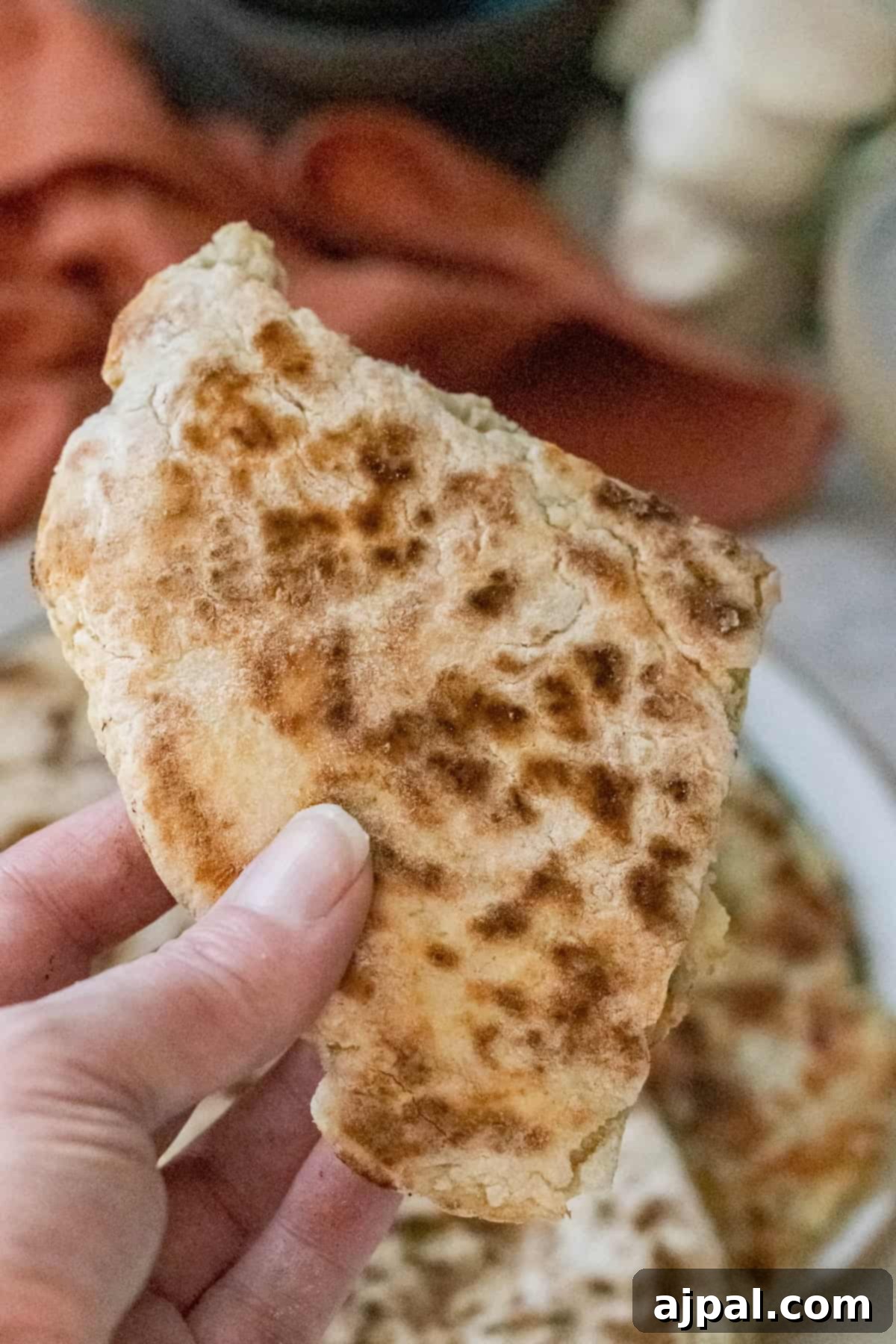 A hand holds a piece of folded, grilled Irish potato bread with a textured surface. In the blurred background, you can spot a plate and cloth, hinting at the traditional skillet method used in the recipe.