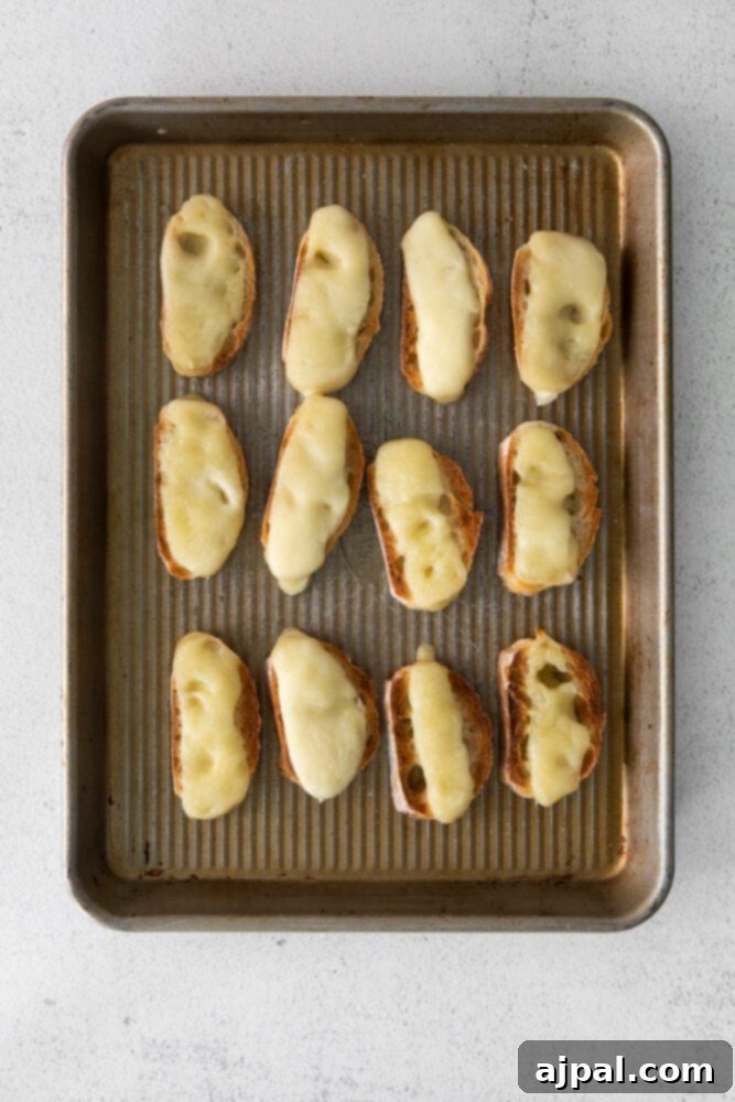 Fresh Caprese Bruschetta 5 Crispy toasted baguette slices topped with melted fresh mozzarella cheese on a parchment-lined baking sheet, part of the Caprese Bruschetta preparation.