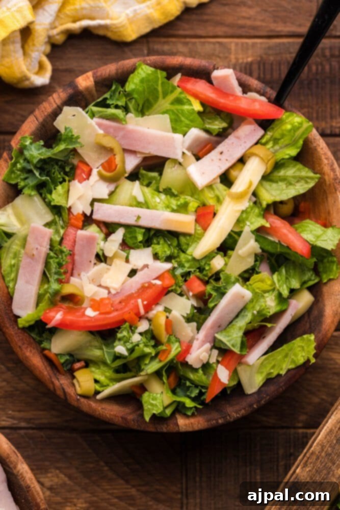 A vibrant Julienne Kale Salad featuring thin strips of kale, romaine, ham, and cheese, drizzled with Mediterranean dressing in a rustic wooden bowl on a table.