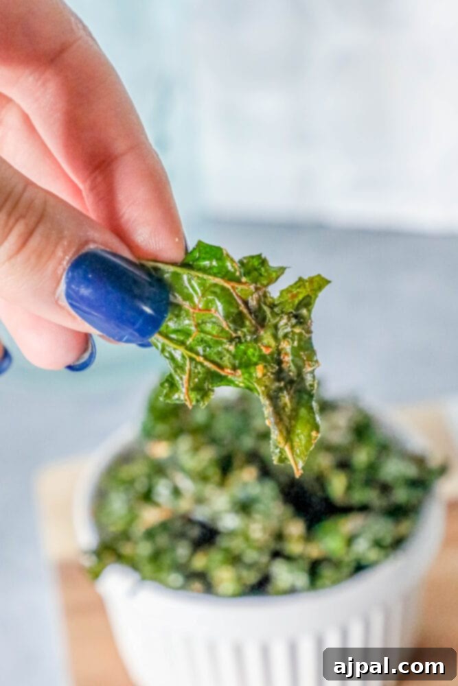 A person's hand holds a small white bowl overflowing with a generous serving of freshly baked, homemade kale chips.