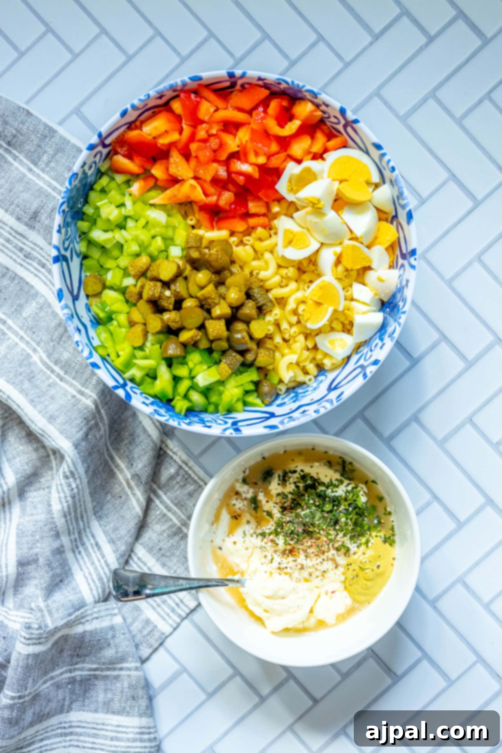 Cooked macaroni in a large mixing bowl, topped with diced gherkins, bell peppers, red onion, celery, and frozen peas, awaiting the creamy dressing