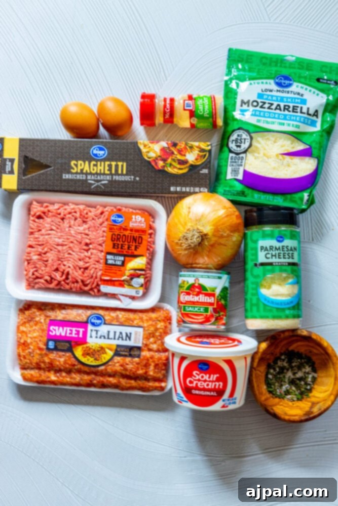 Assortment of spaghetti pie ingredients including pasta, eggs, garlic powder, mozzarella, onion, pasta sauce, sour cream, parmesan cheese, and Italian herbs arranged on a table.