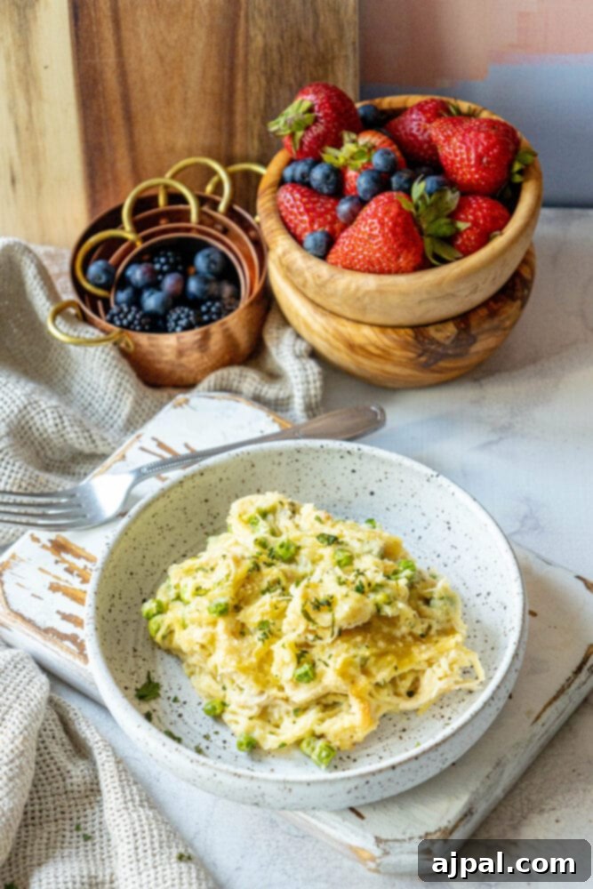 A close-up of a serving of baked chicken tetrazzini noodle casserole with peas on a white plate, showing the creamy texture and golden cheese crust, with a bowl of fresh berries in the background.