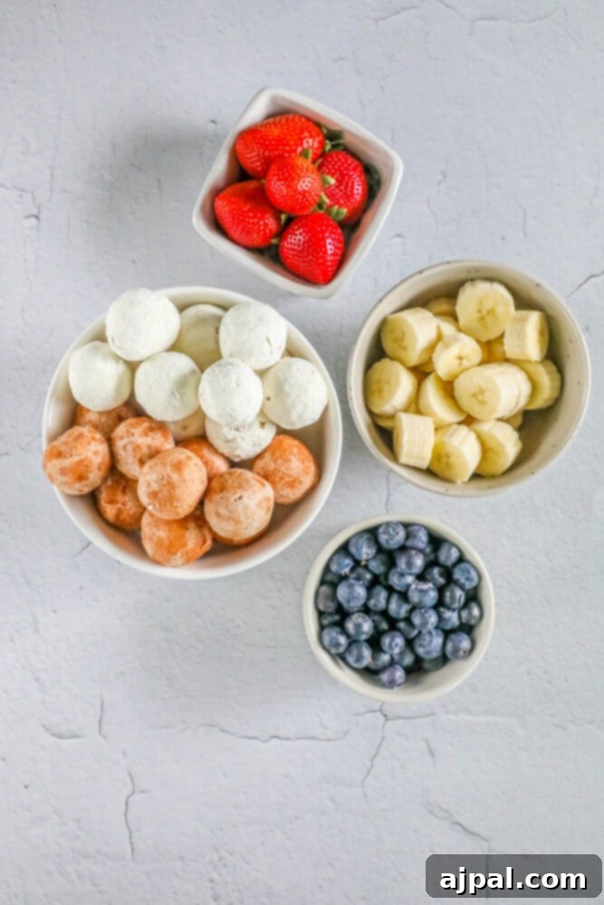 Red, White, and Blue Dessert Skewers 5 A colorful array of ingredients for dessert kabobs, including bowls of fresh strawberries, blueberries, sliced bananas, and donut holes, ready for assembly on a clean surface.