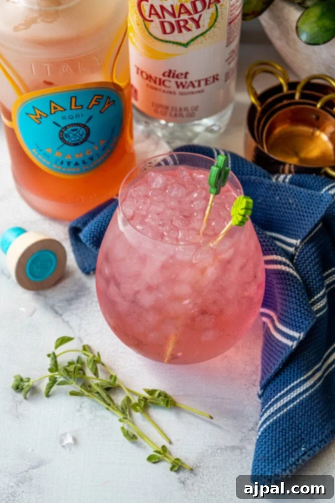 Sunset Sip Blood Orange Gin Tonic 2 Pink gin and tonic in a large glass with ice, in front of a bottle of Malfy Gin Con Arancia.
