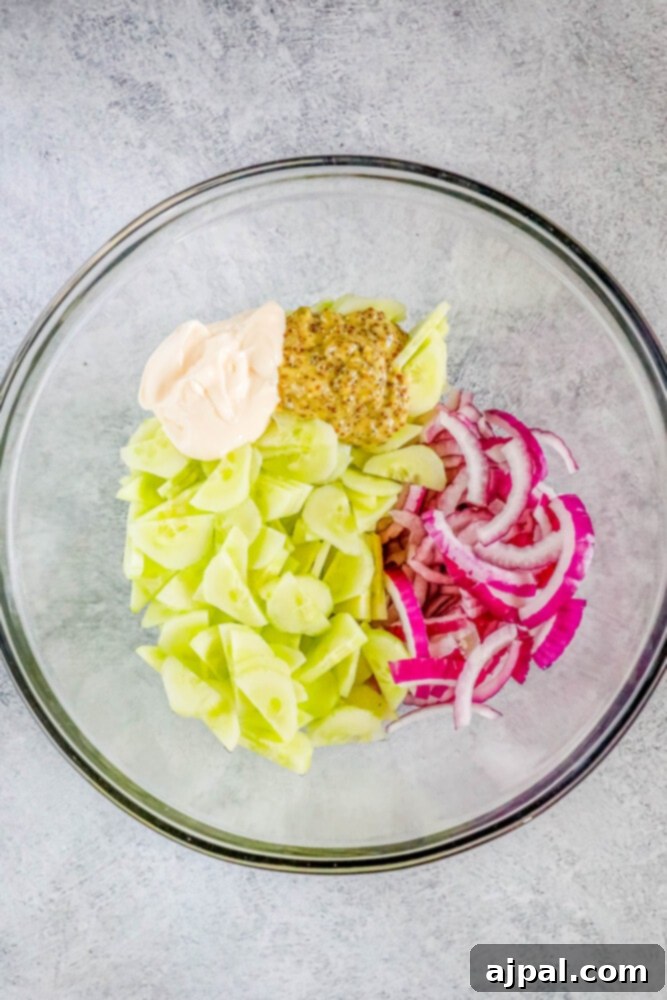 Cool Creamy Cucumber Salad 7 A close-up of sliced cucumbers and red onions in a white bowl, with dollops of mayonnaise and whole grain mustard added, ready for mixing to create a creamy cucumber salad.