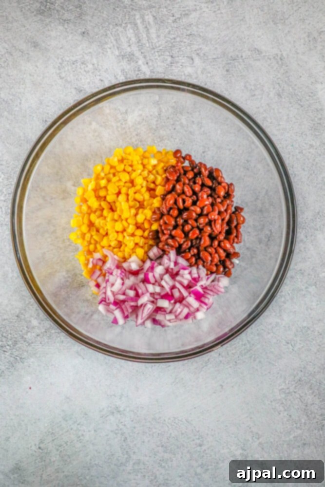 A glass bowl containing corn, diced red onion, and black beans, showcasing the initial mix for salsa.