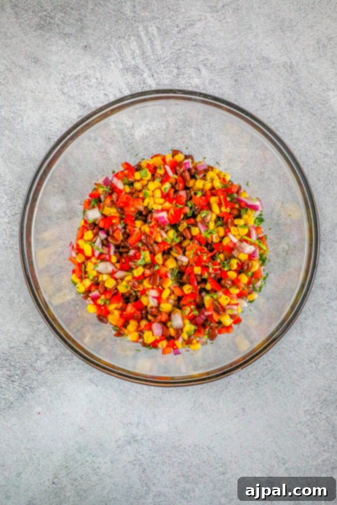 Diced bell pepper, corn, beans, and diced red onion combined in a clear glass bowl for salsa.