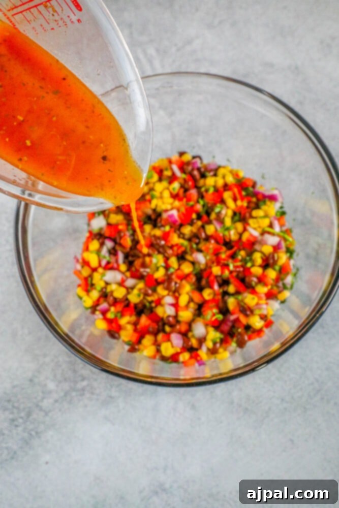 Lime juice and herbs being poured into a glass bowl filled with diced peppers, beans, onions, and corn for salsa dressing.