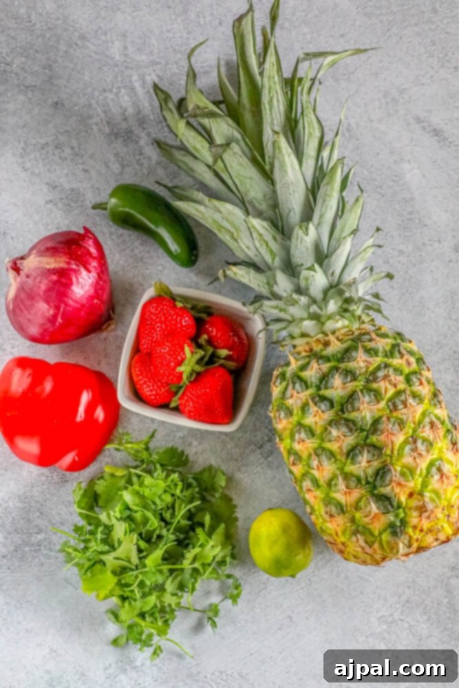 Array of fresh ingredients including pineapple, jalapeno, strawberries, red onion, cilantro, lime, and red bell pepper on a rustic wooden table, ready for chopping.