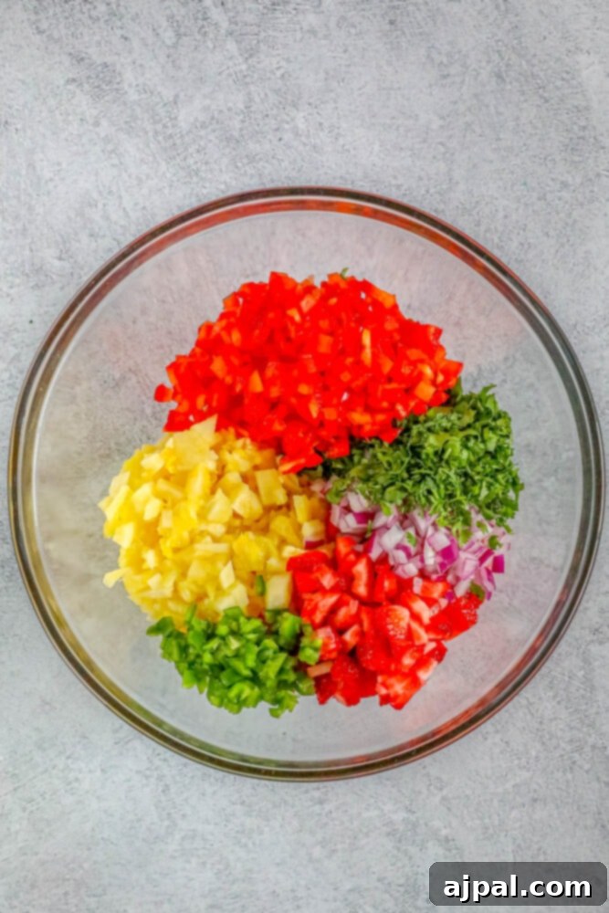 Diced pineapple, jalapeno, strawberries, red onion, cilantro, and red bell pepper in a clear mixing bowl on a wooden table, showcasing their vibrant colors.
