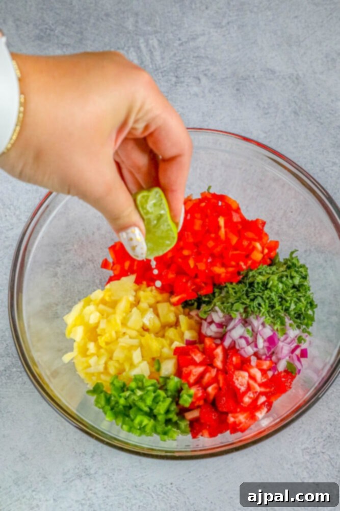 A hand squeezing fresh lime juice over a bowl of pineapple, jalapeno, strawberries, red onion, cilantro, and red bell pepper on a wooden table.
