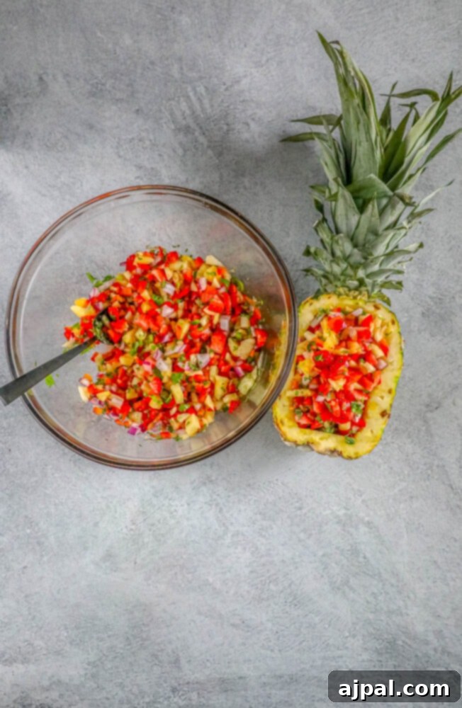 A glass bowl of strawberry pineapple salsa with a spoon, being scooped into an empty pineapple hull, preparing for a festive presentation.