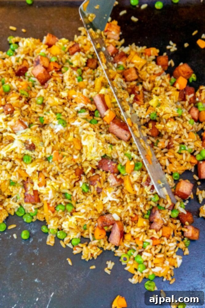 Effortless Spam Fried Rice 10 picture of spatula mixing sauces and fried eggs into rice, peas, onion with fried spam frying on a griddle