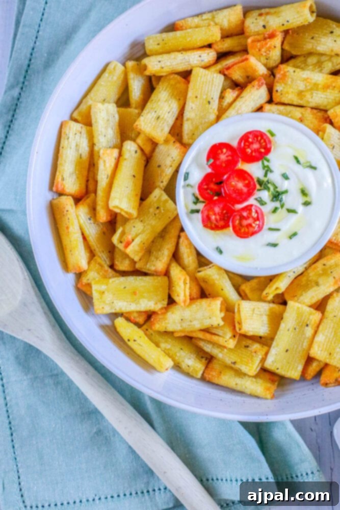 A bowl of golden air-fried pasta chips served alongside a creamy feta dip with fresh tomatoes, inviting a delicious snack.