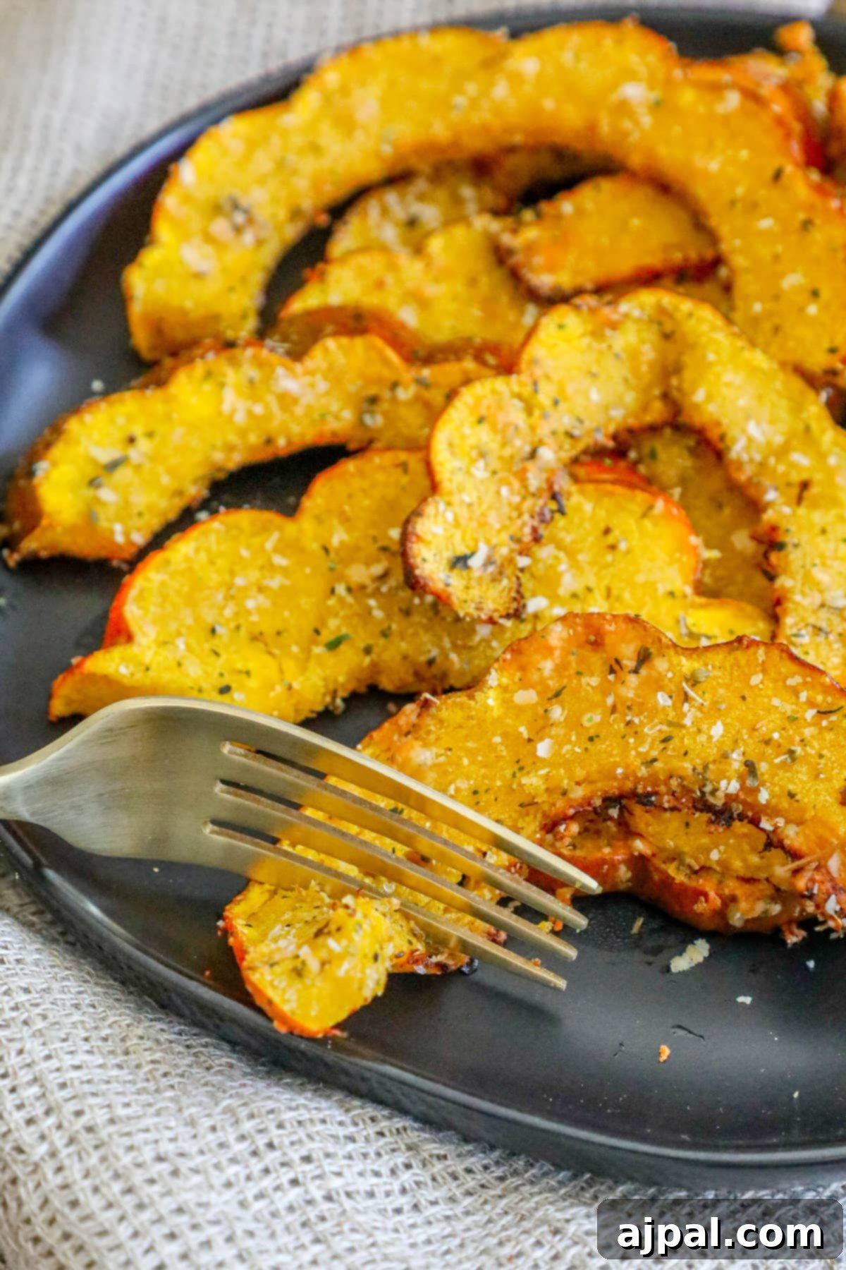 Picture of roasted acorn squash slices seasoned with herbs and spices on a black plate, garnished with a sprig of fresh herbs and a fork.