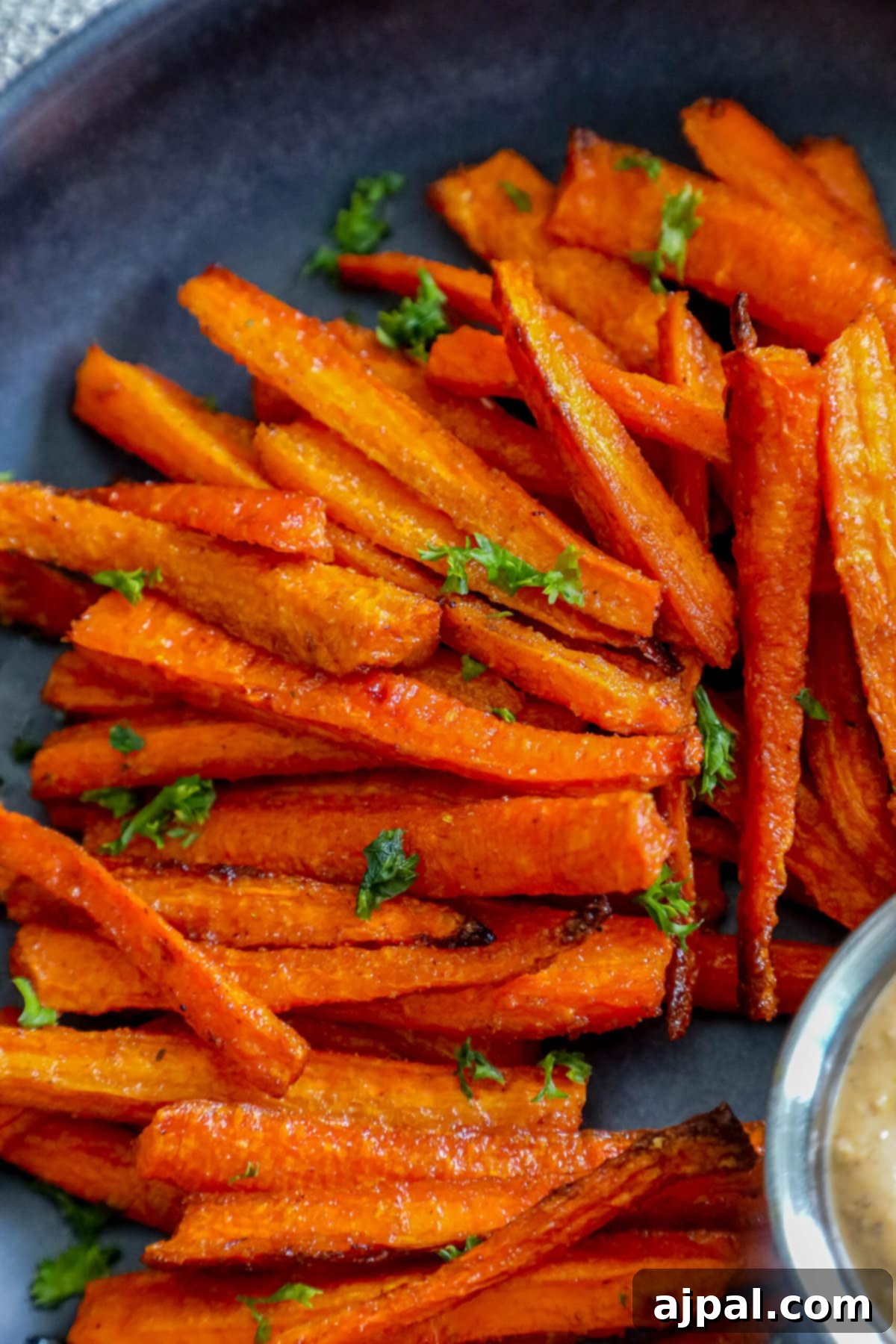 Crispy baked carrot fries garnished with fresh parsley on a black plate, with a side of dipping sauce.