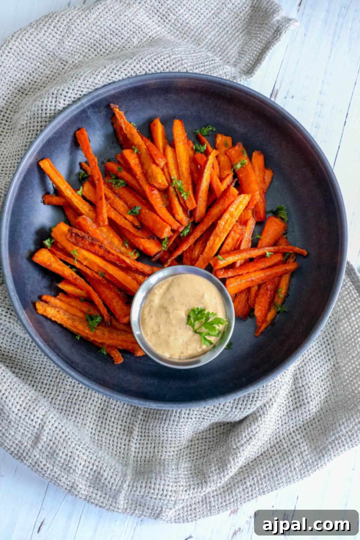 Plate of baked carrot fries with fresh parsley and a cup of dipping sauce on a black surface.