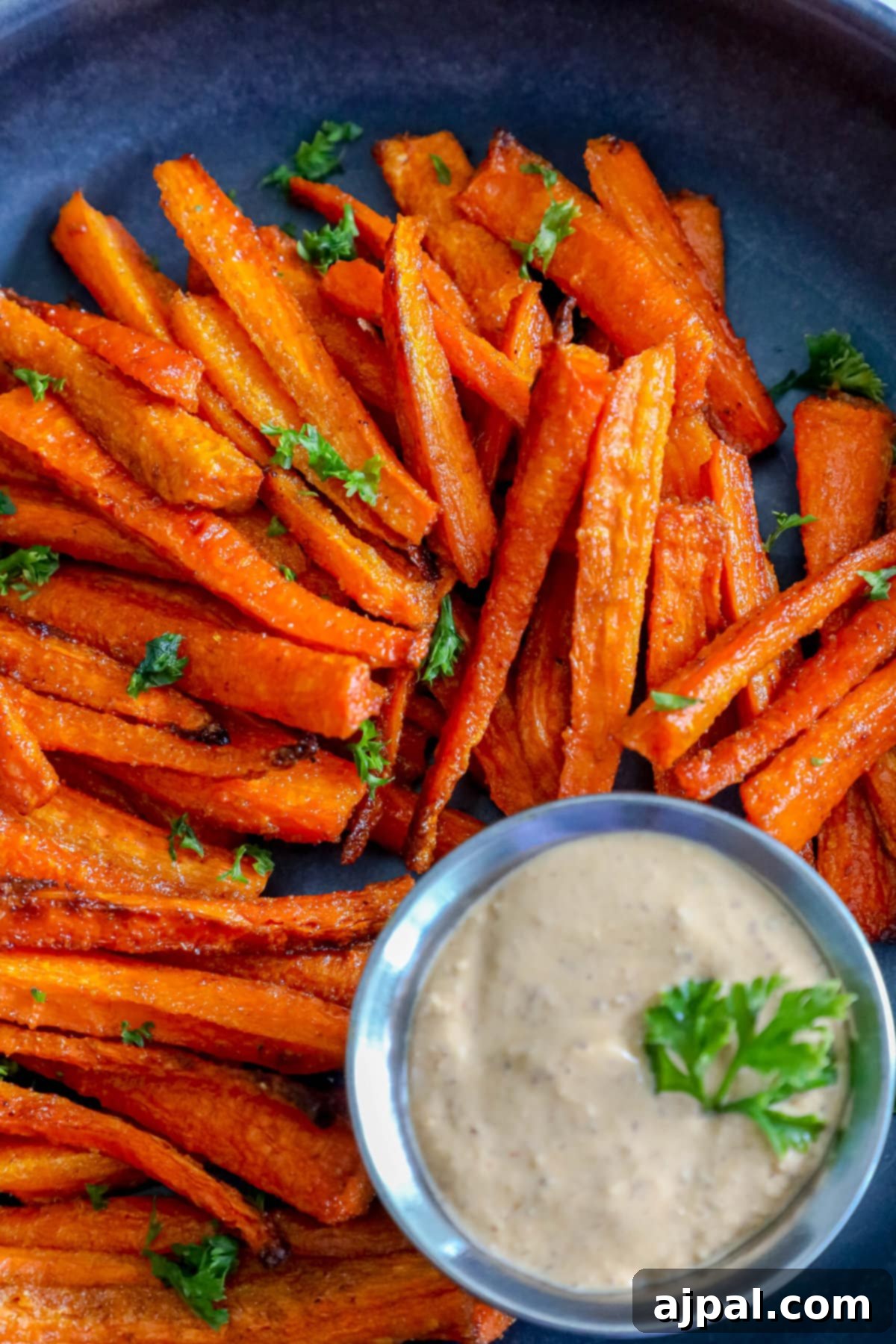 picture of carrot fries with parsley chopped on top on a black plate with a cup of sauce on the side
