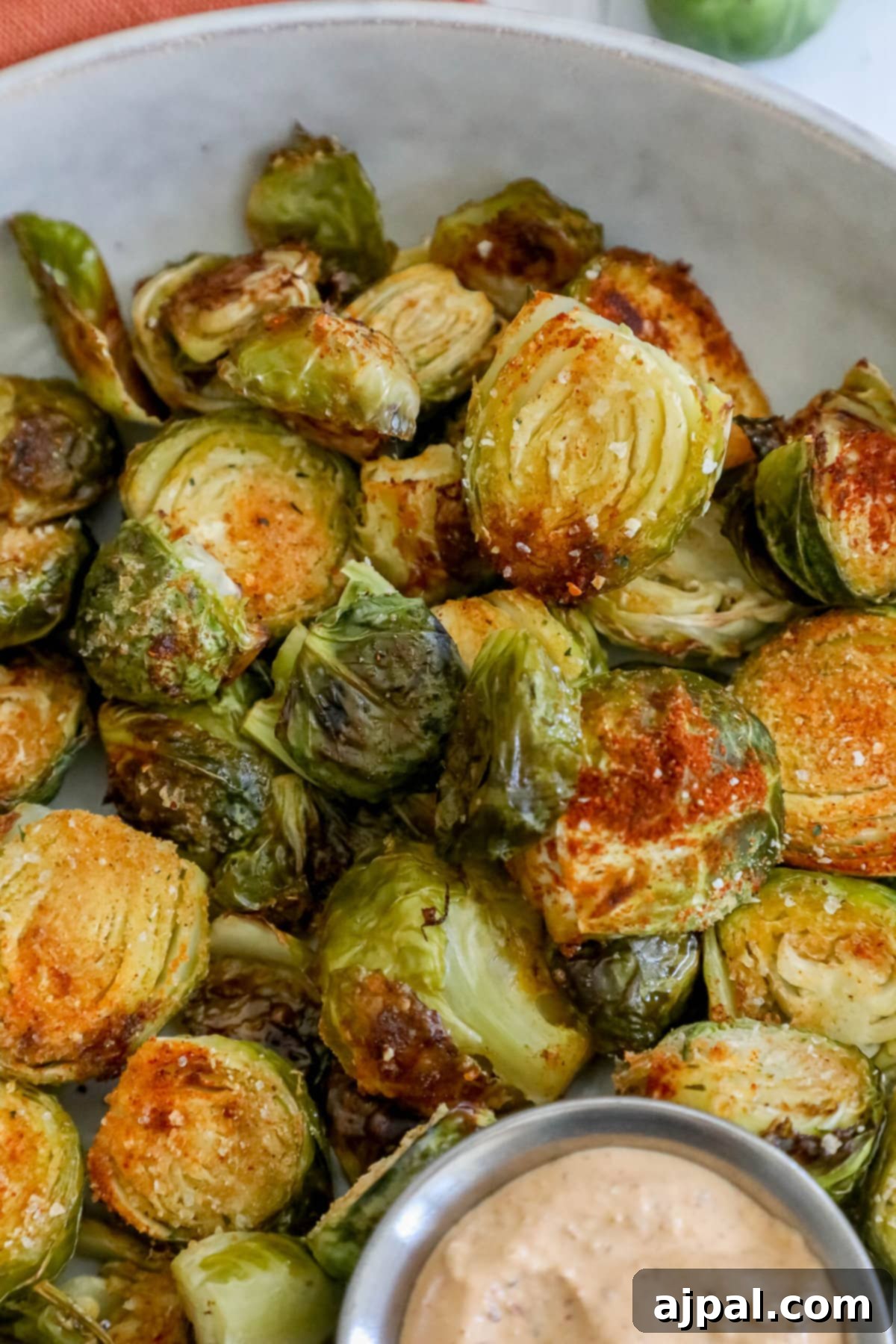 Crispy Garlic Roasted Brussels 11 A white bowl showcasing garlic roasted Brussels sprouts, perfectly seasoned and cooked, with a small cup of sauce.