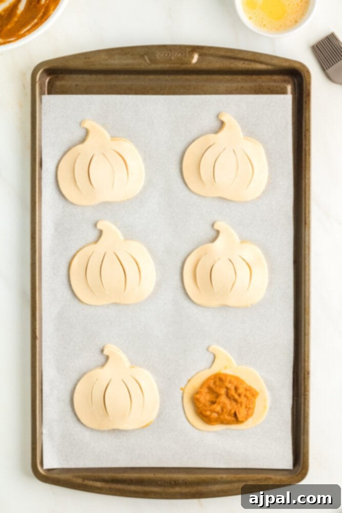 Autumn Pumpkin Pockets 10 Two pumpkin-shaped pie dough pieces, one with pumpkin filling, arranged on a parchment-lined baking sheet.