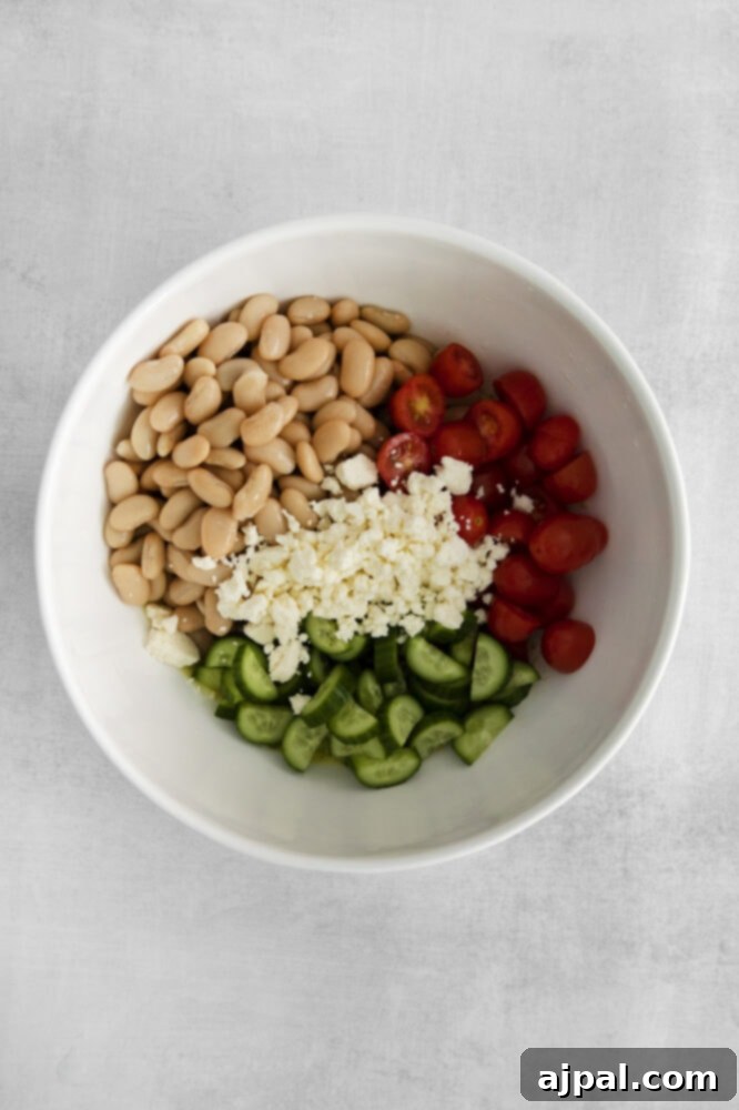 Close-up of cannellini beans, chopped cucumbers, sliced cherry tomatoes, and crumbled feta cheese prepared in a bowl on a wooden table.