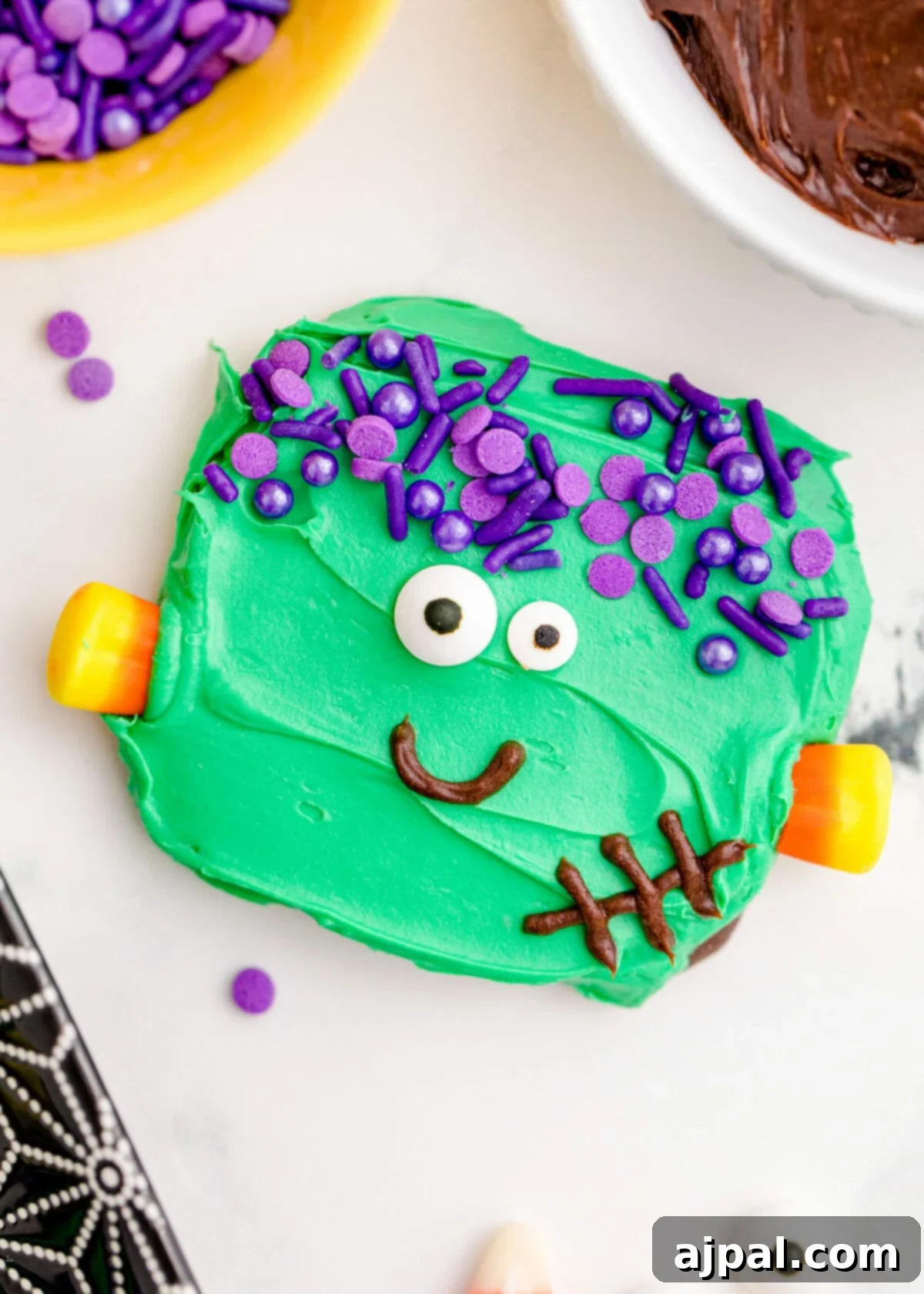 Frankenstein Cookies 2 A close-up shot of an adorable Frankenstein cookie with green icing, purple sprinkle hair, candy corn neck bolts, expressive candy eyes, and a chocolate icing smile, presented on a cooling rack.