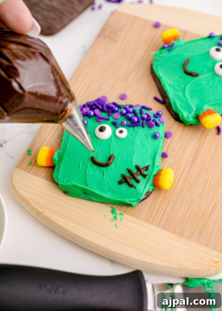 Spooky Frankenstein Cookies 11 A hand meticulously piping a chocolate icing smile onto a green Frankenstein cookie, adding a cheerful touch to the monster's face.
